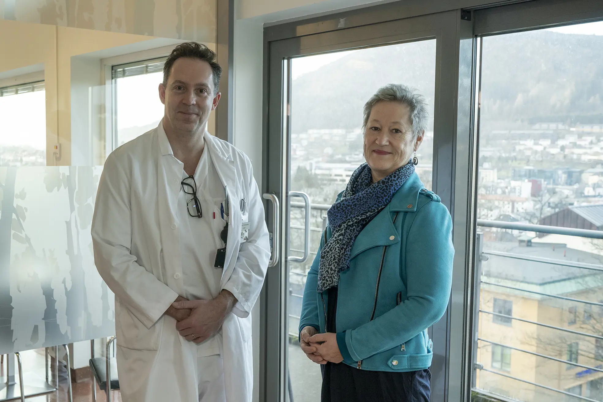 Lege kledd i hvitt og eldre kvinne står foran glassdør. Foto