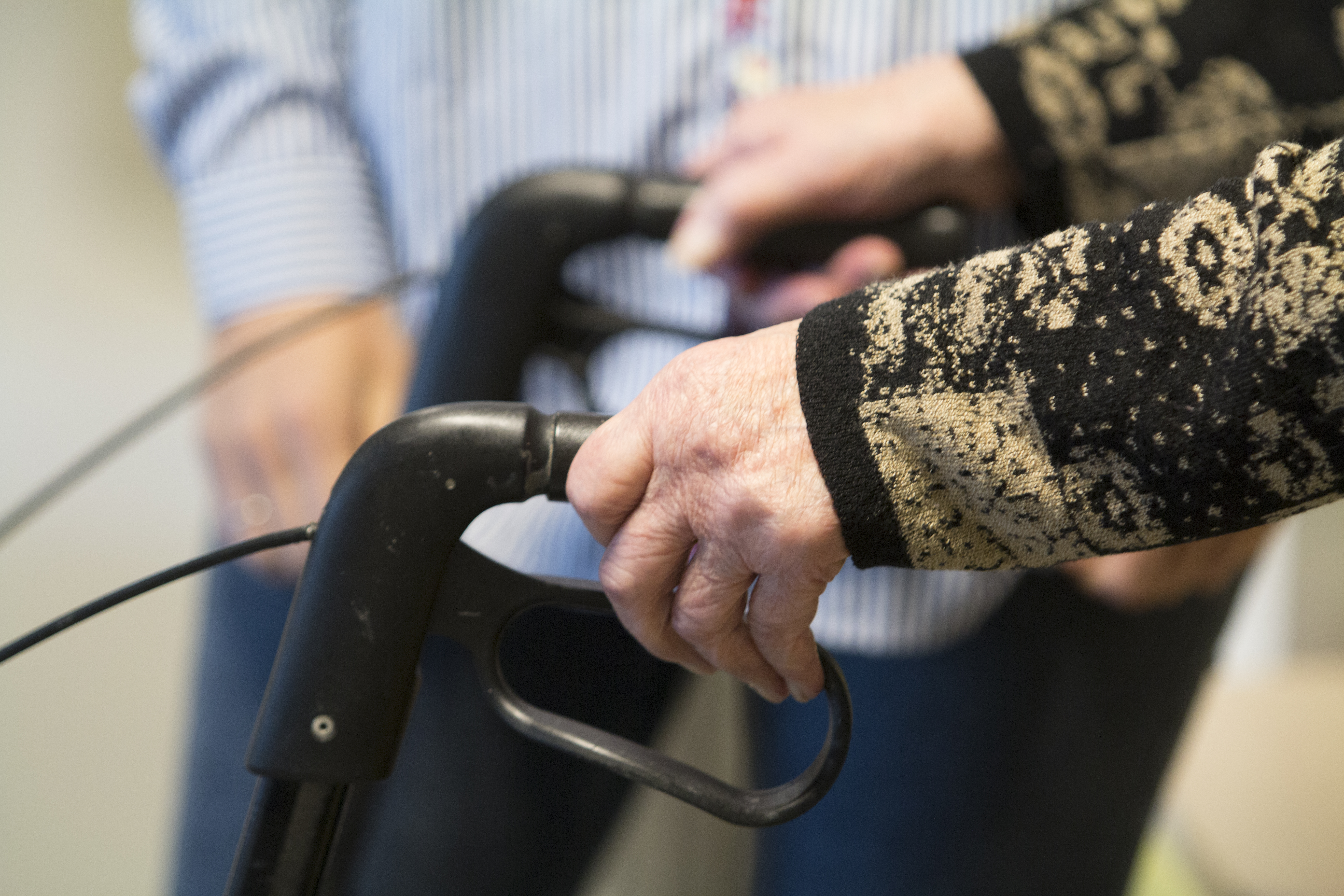 Hendene til eldre person held på gåstolhandtak med person i bakgrunnen.
