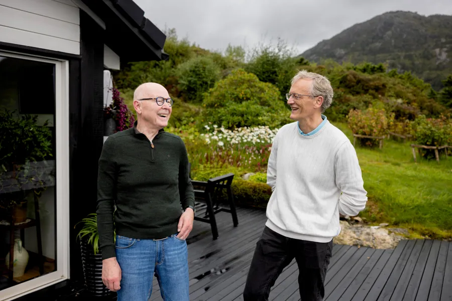 Ståle Myklebust og Halfdan Sørbye som smiler og snakker på et treterrasse utenfor et hus, med grønne åser og overskyet himmel i bakgrunnen.