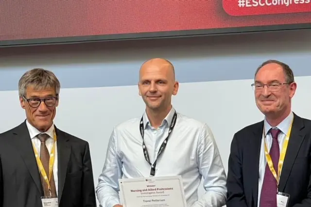 Gruppebilde av tre menn, mann i midten held eit diplom.