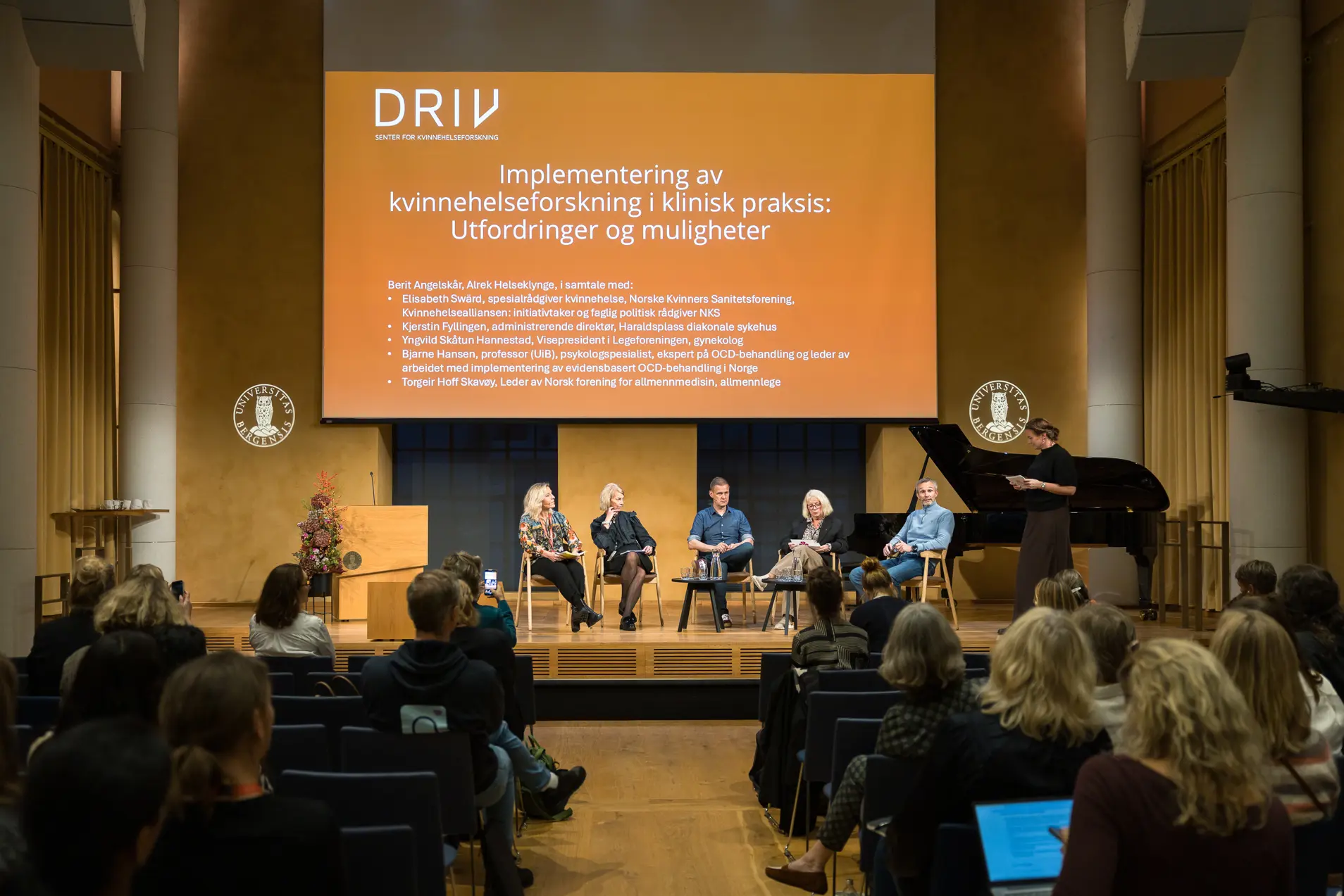 En gruppe mennesker som sitter i et rom med en scene og en stor skjerm