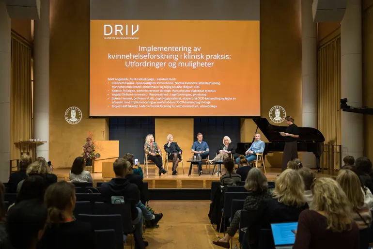 En gruppe mennesker som sitter i et rom med en scene og en stor skjerm