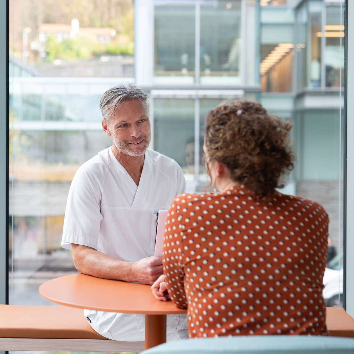 Mannlig sykepleier og kvinne sitter ved et bord ved vindu.