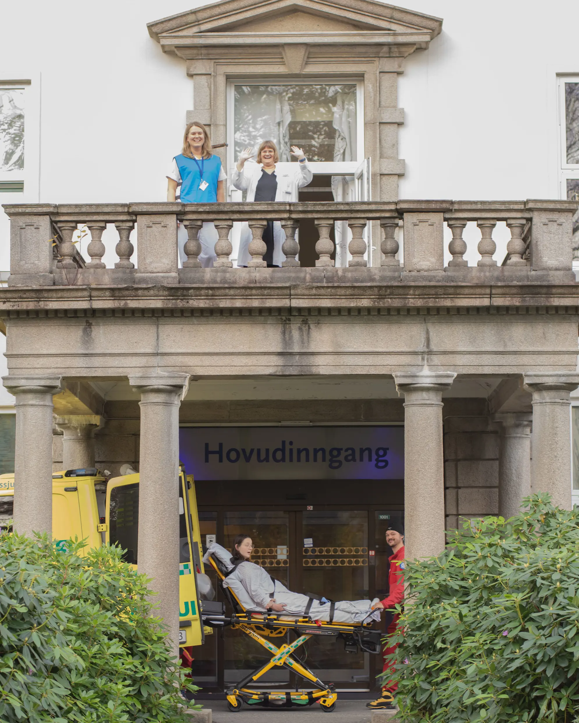 To kvinner står på balkong, under balkongen flyttast ei kvinne på ambulansebåre inn i ambulanse.