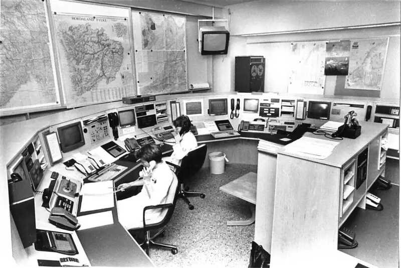 Et historisk foto av to personer som sitter ved en sentral med mange skjermer