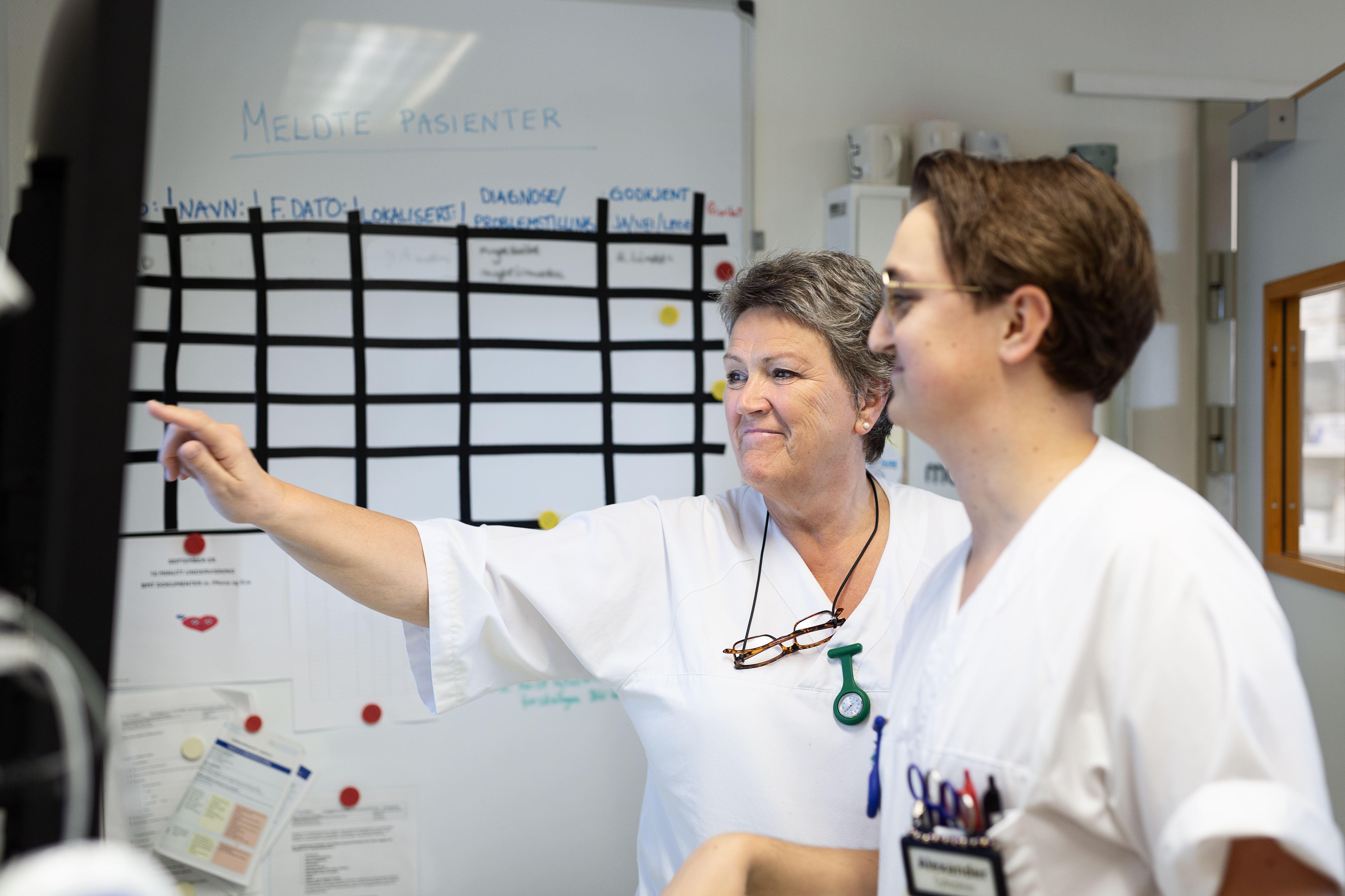 Helsepersonell i hvitt på vaktrom diskuterer foran en skjerm