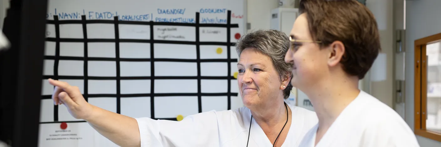 Helsepersonell i hvitt på vaktrom diskuterer foran en skjerm