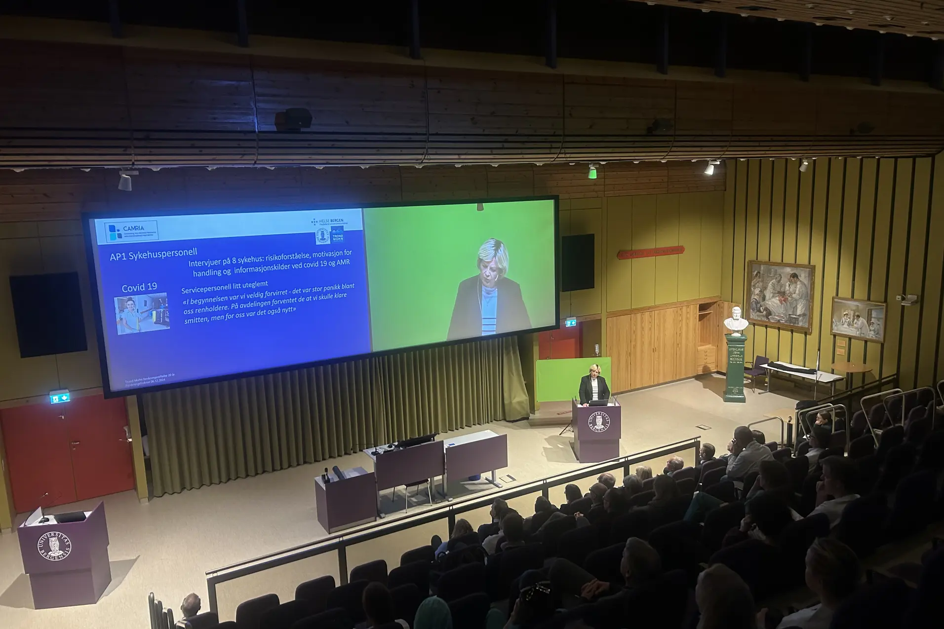 Et stort rom med en stor skjerm og en gruppe mennesker som sitter i stoler