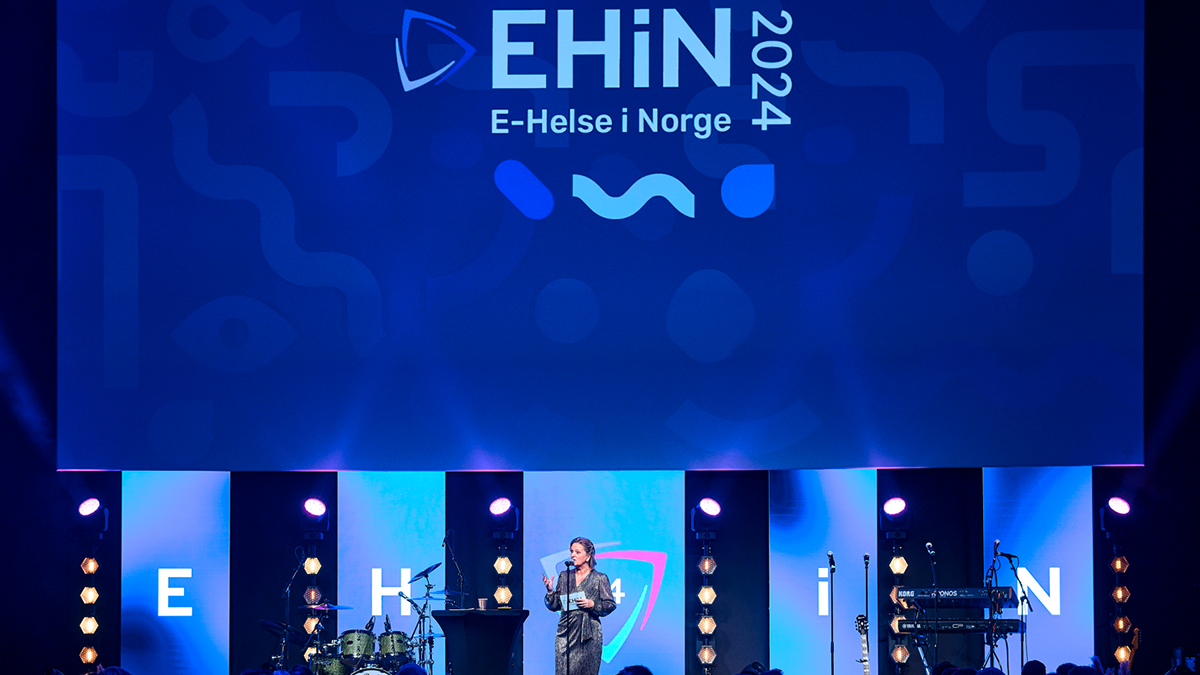 En person på en scene med mikrofon og en stor skjerm bak seg