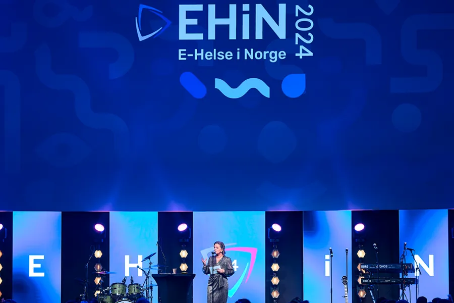 En person på en scene med mikrofon og en stor skjerm bak seg